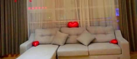 Una sala de estar moderna con un sofá modular, una mesa de centro y un ventanal con cortinas traslúcidas y adornos con forma de corazón.