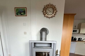 A room with a wood-burning stove, a wall clock, and a kitchen area. - Apartment in Frankenau with swimming pool (Frankenau)