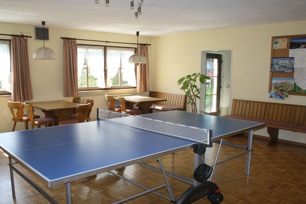 Une pièce avec une table de ping-pong, un plancher en bois, une table à manger avec des chaises, un banc et un tableau d’affichage avec des feuilles de papier.