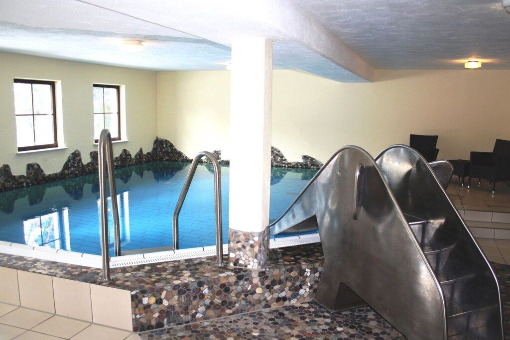 An indoor swimming pool with a stone-tiled edge, metal steps, and a view of the sky through windows.