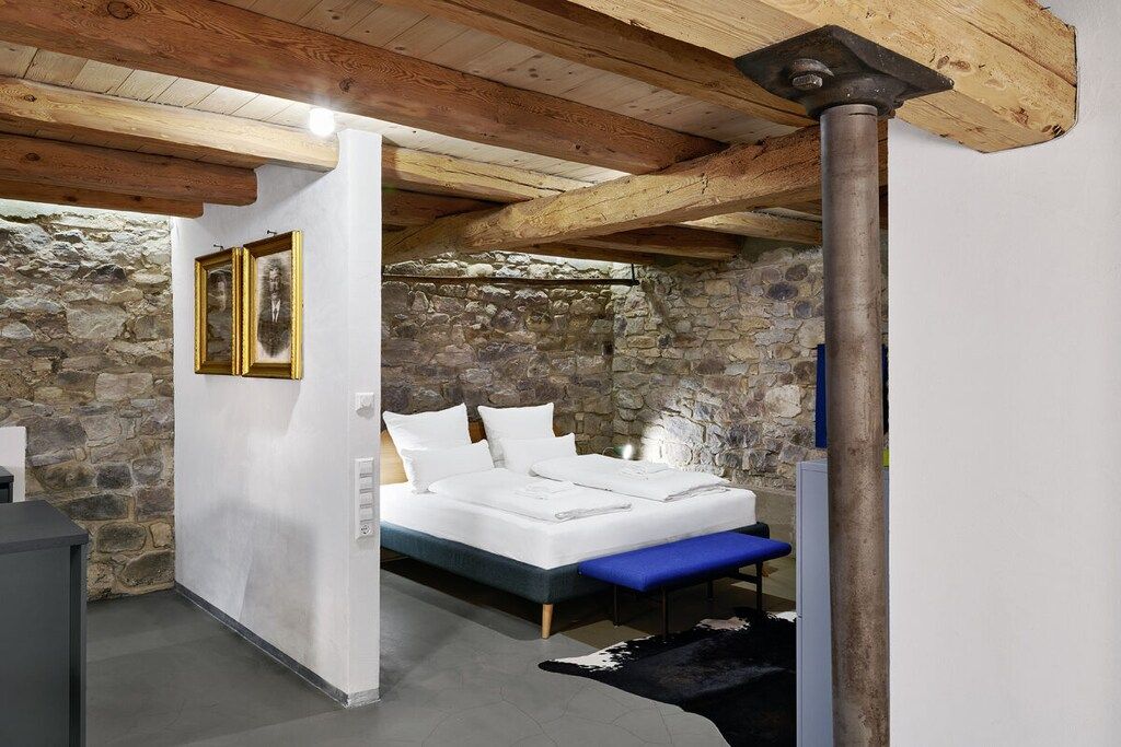 Une chambre avec un mur en pierre, un lit avec une literie blanche, un banc bleu et un plafond en bois.