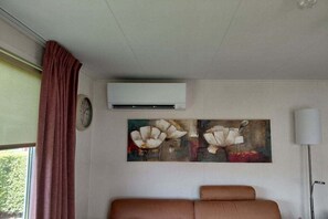 A room with a brown sofa, a clock, an air conditioning unit, and a painting of flowers on the wall.