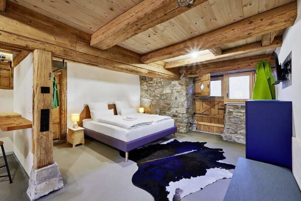 A bedroom with a stone wall, wooden beams, a bed, a nightstand, a TV, and a window.