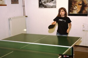 A person playing table tennis in a room with a radiator, posters on the wall, and a table tennis table. - 2 Houses on the edge of the forest (Leutenbach)