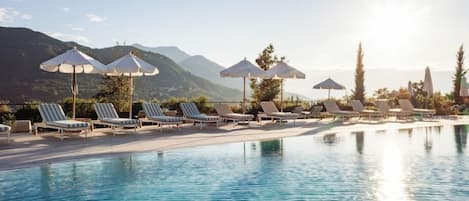 Zona de piscina con sillones de descanso y sombrillas, cielo despejado y montañas al fondo.