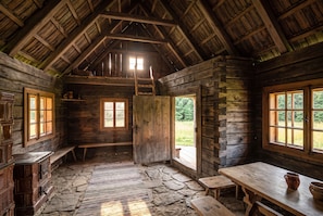 A rustic cabin interior with a stone floor, wooden walls, a loft area, and large windows.
