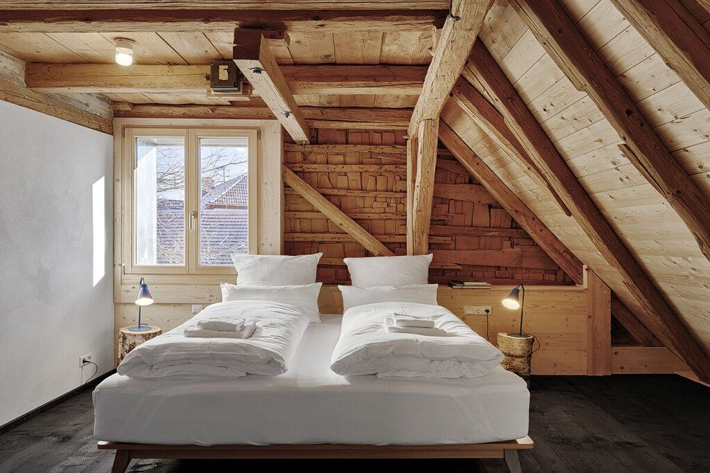 A room with a wooden ceiling, a bed with white linens, a window with a view, and a small table with a lamp.