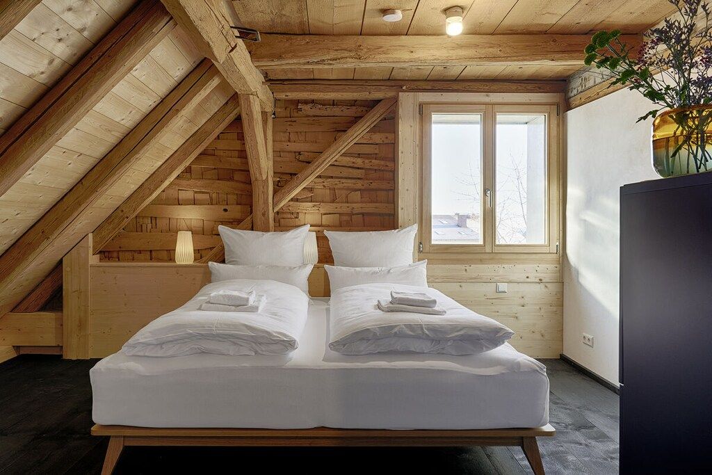 Une chambre avec un plafond en pente en bois, un lit avec du linge de lit blanc, une fenêtre donnant sur un beau paysage et un vase rempli de fleurs posé sur une commode.
