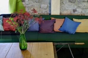 Un sofá verde con varios cojines de diferentes colores, una mesa de madera con un jarrón de flores, y una pared de piedra al fondo.
