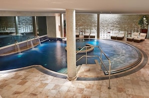 An indoor swimming pool with a circular design, surrounded by lounge chairs and a stone wall.