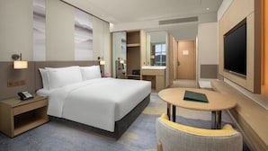 Quarto de hotel com cama, mesa de cabeceira, luminária, TV e uma pequena mesa redonda com uma cadeira.