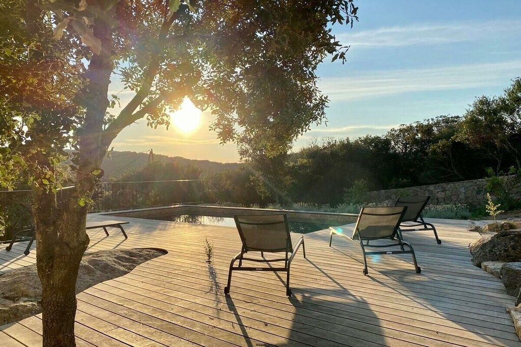 A wooden deck with two chairs and a pool, surrounded by trees and a stone wall.