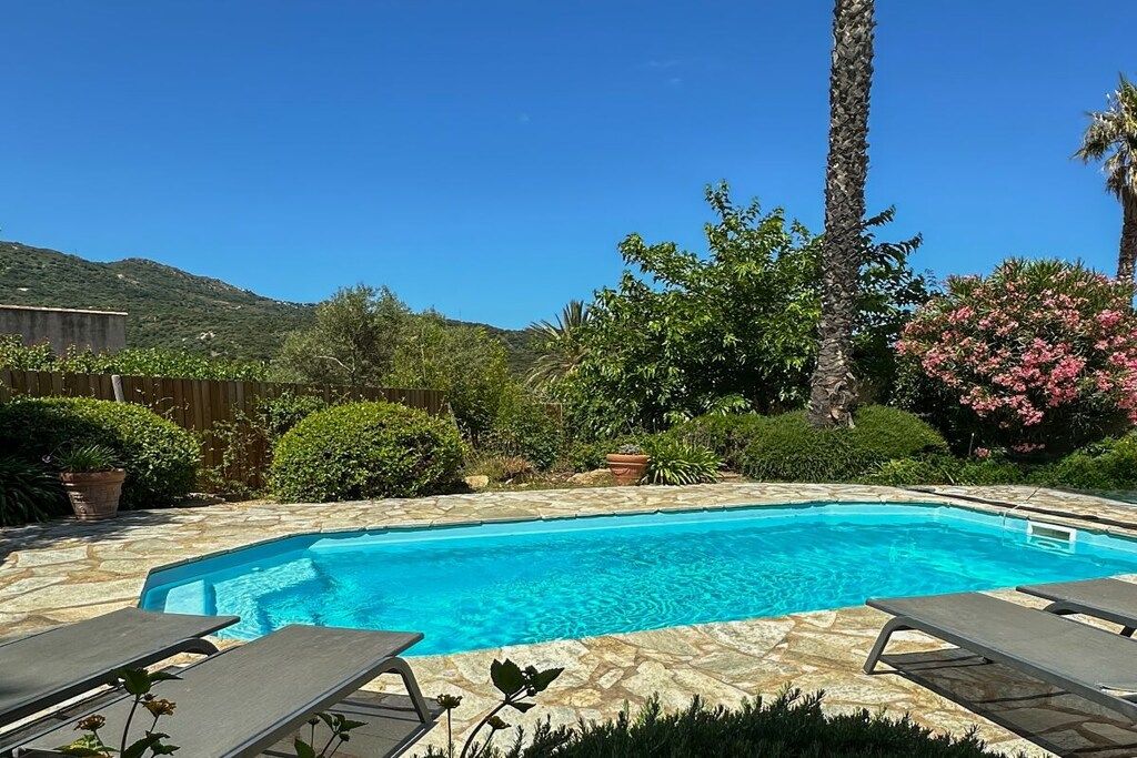 A clear blue swimming pool with stone tiles, surrounded by lush greenery and a wooden fence.