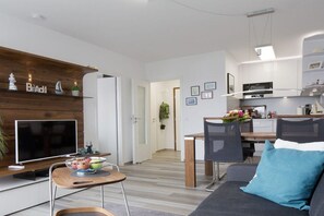 Ein modernes Wohnzimmer mit Flachbildfernseher, einer Holzregal mit Dekoration, einem Essbereich mit Tisch und Stühlen sowie einer Küche im Hintergrund.