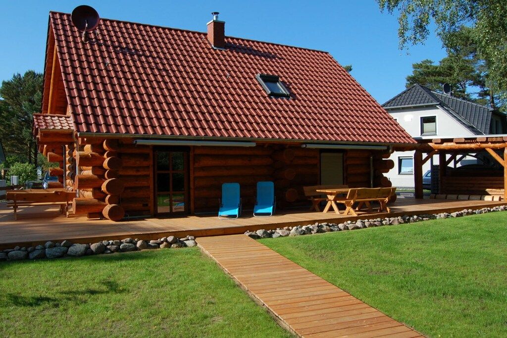 Une cabane en rondins avec un toit en tuiles, une terrasse en bois et un chemin de pierre menant à l’entrée.