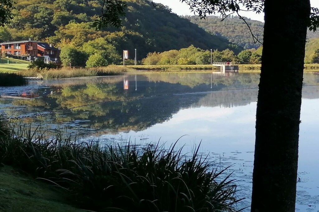 Un lago tranquilo con el reflejo de un edificio y árboles, rodeado de vegetación y colinas.
