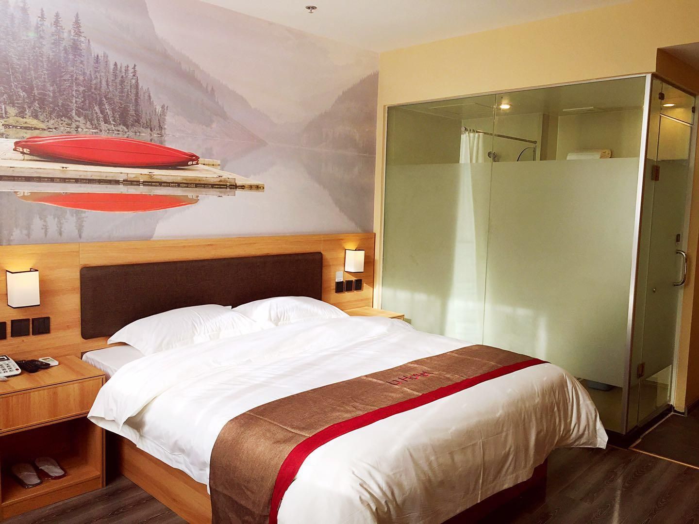 A hotel room with a bed, a nightstand with a phone, a glass shower enclosure, and a mural on the wall.