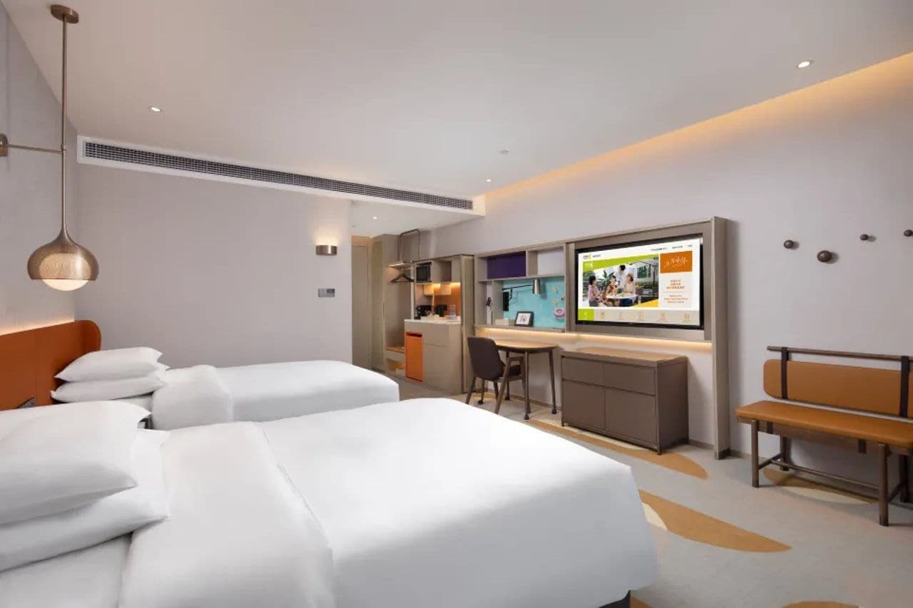Habitación de hotel con dos camas, un escritorio, una banca y un televisor mostrando una revista.