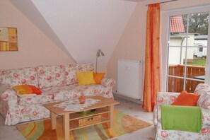 A cozy living room with a floral sofa, a wooden coffee table, and a view of the outdoors through a window with orange curtains. - Holiday apartment with 2 bedrooms (Kölpinsee)