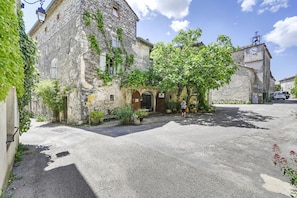 Ein Steinhaus mit einem zentralen Eingang, einem gepflasterten Innenhof und einem geparkten Fahrzeug im Hintergrund.