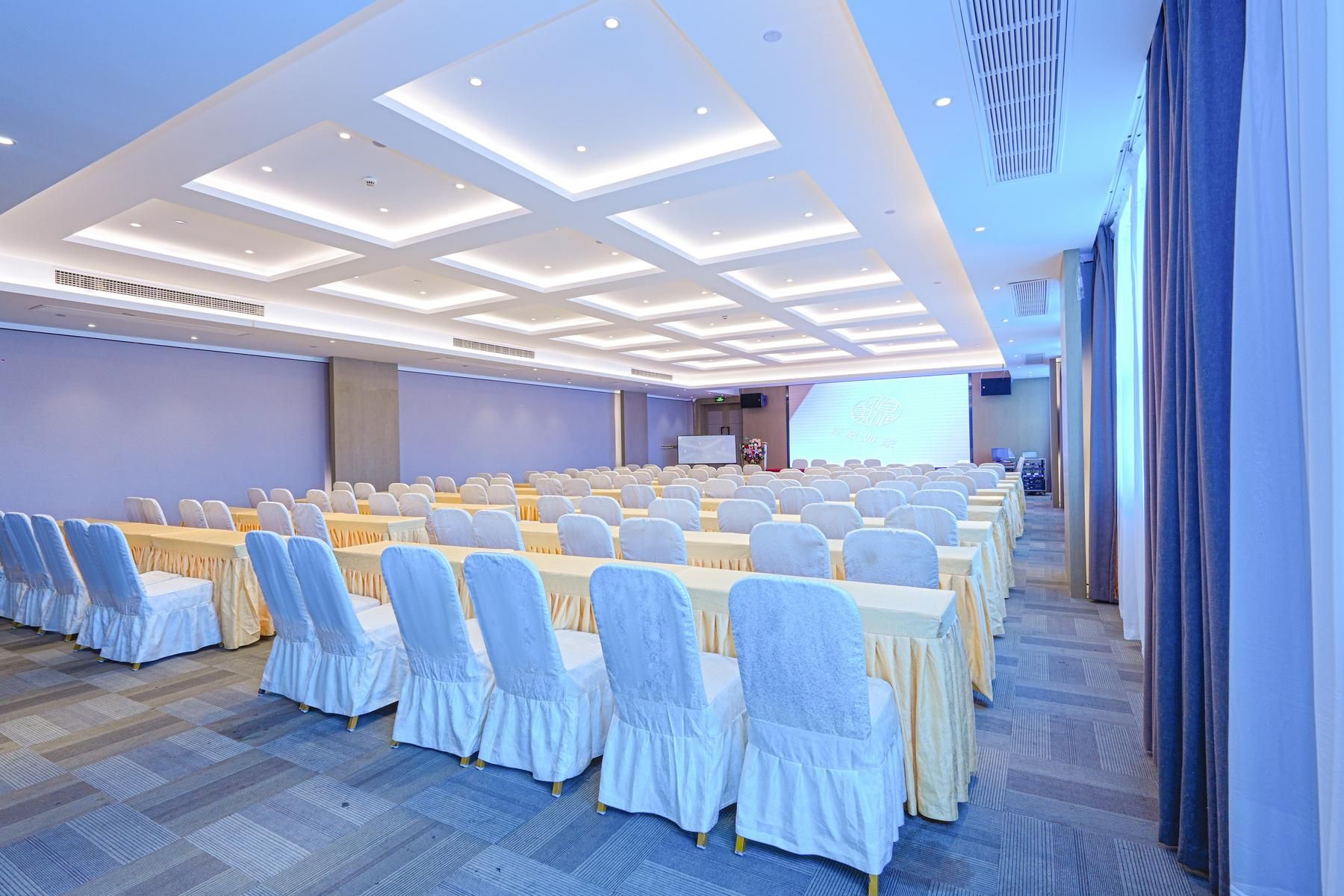 Un salón de conferencias con filas de mesas y sillas, una pantalla grande y un escenario.