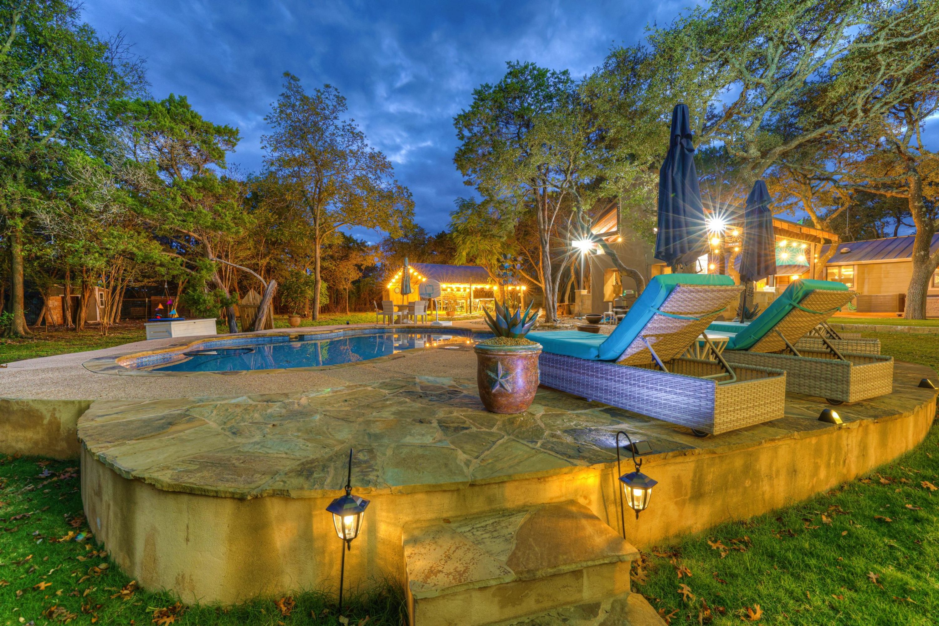 Una piscina con sillones de descanso y sombrillas, rodeada de árboles y un edificio con luces.