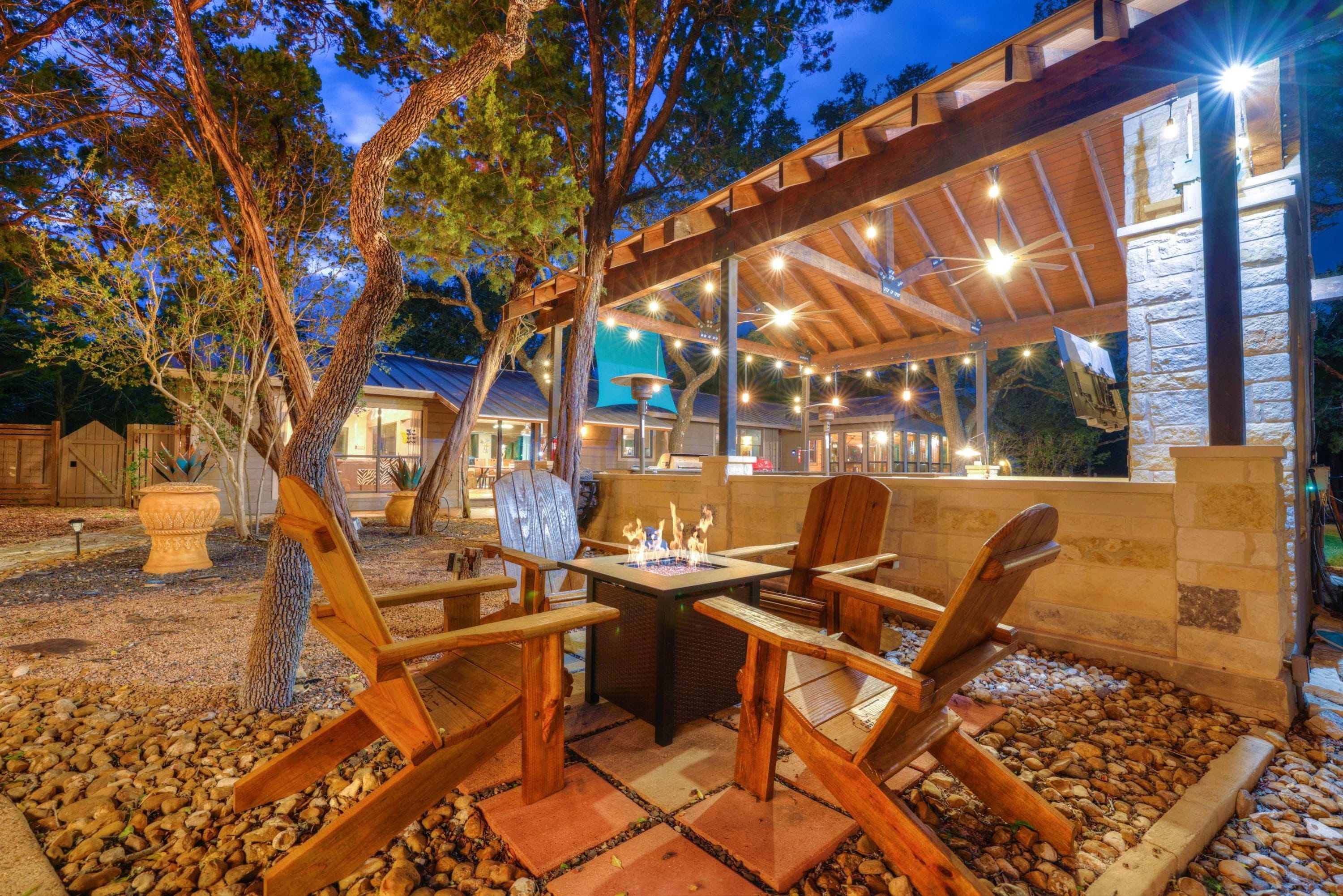 An outdoor patio with wooden furniture and a fire feature, surrounded by trees and a stone wall.