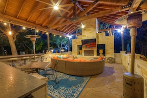 Un patio exterior con suelo de piedra, una zona de estar circular, una chimenea y luces colgantes.