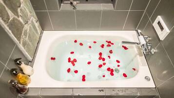 A bathtub with rose petals floating on the water, surrounded by grey tiles and a stone wall.