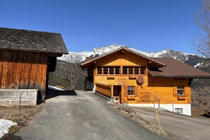 Ein hölzernes Gebäude mit geneigtem Dach, einem sichtbaren Eingang und freiem Blick auf schneebedeckte Berge im Hintergrund.