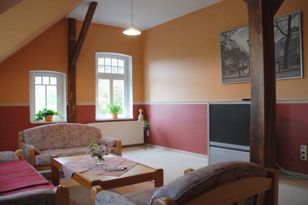 Een kamer met een schuine kap, een zithoek, een tafel met een vaas en twee ramen met planten.