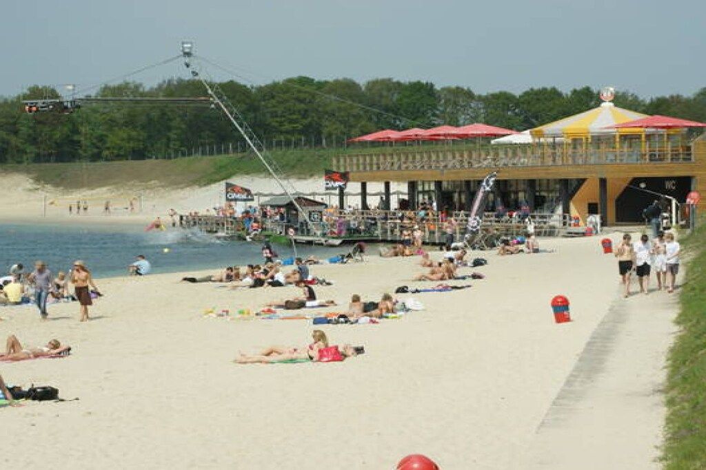 Een strand met mensen die zonnebaden en wandelen, een pier met een boot, en een gebouw met een geel dak en rode parasols.