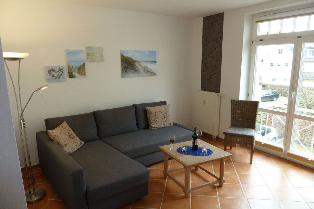 Ein Wohnzimmer mit einer Couch, einem Tisch mit Flasche und Glas sowie einem Sessel. An den Wänden befinden sich Kunstwerke und ein Blick durch eine Glastür.