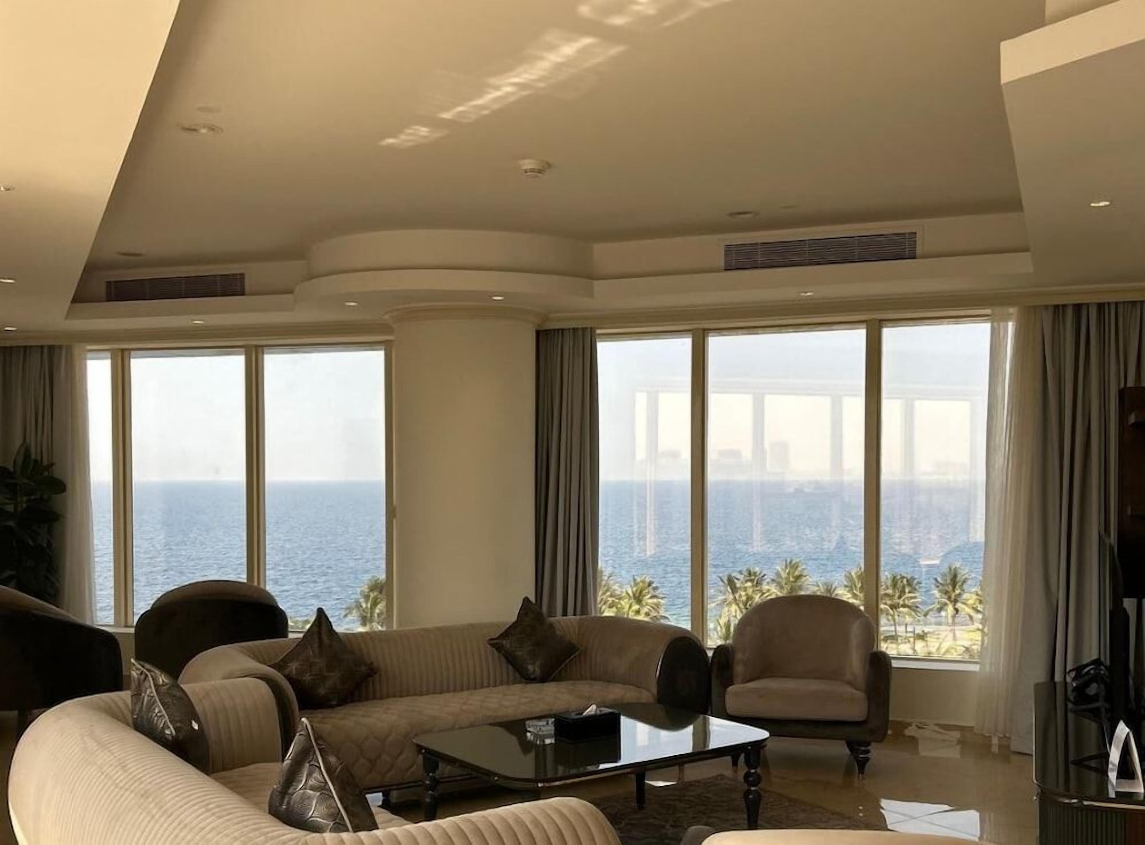 Een ruime woonkamer met een groot raam dat uitzicht biedt op de oceaan en palmbomen, een comfortabele zithoek en een salontafel.