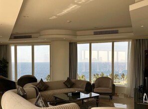 A spacious living room with a large window offering a view of the ocean and palm trees, a comfortable sofa set, and a coffee table. - Seafront Luxury Suites (Jeddah)