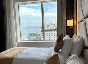 A hotel room with a bed, a nightstand, a lamp, and a view of the sea and buildings.