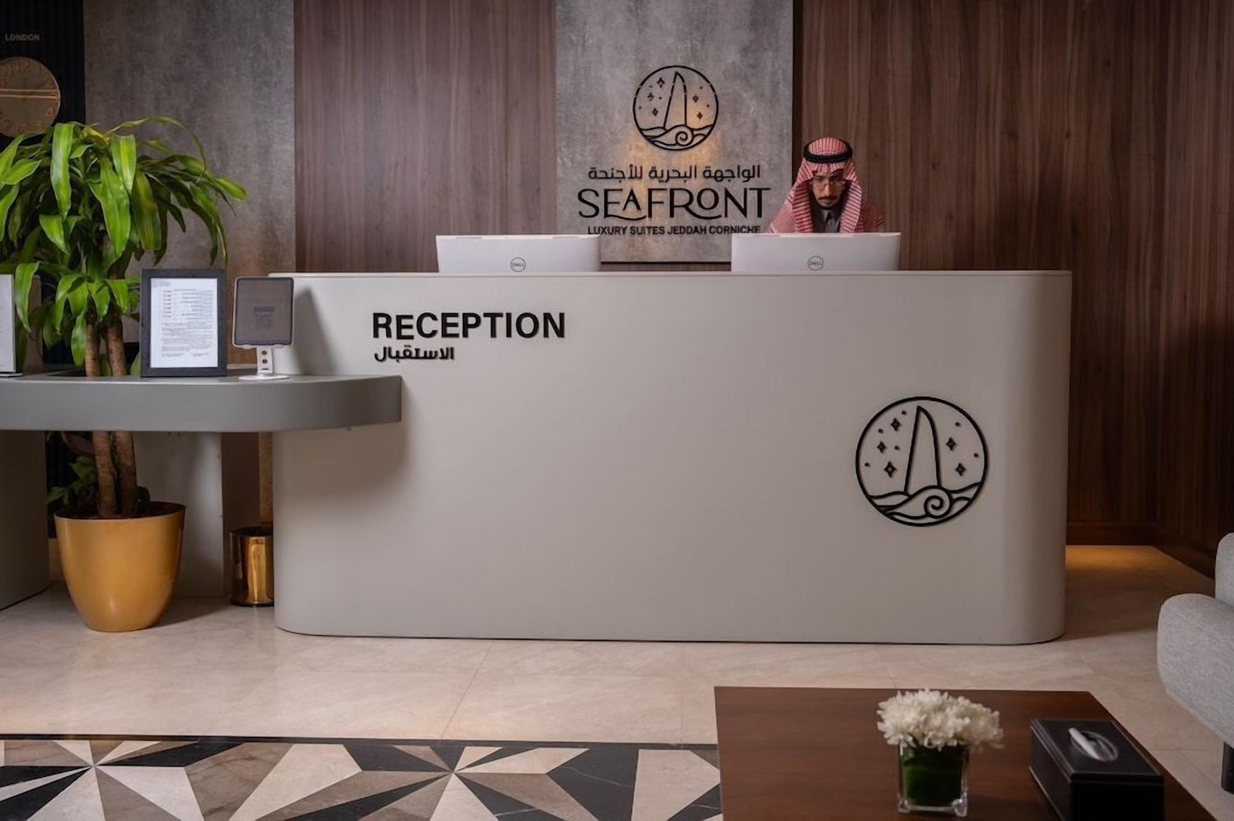 Un mostrador de recepción con un cartel que indica 'RECEPTION' y 'SEAFRONT' en varios idiomas, una computadora portátil, una planta y un documento enmarcado.