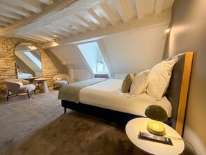 A bedroom with a large bed, a round bedside table, a chair, and a stone wall. - Villa Ursuline: Charm and Comfort in the Heart of the City (Bayeux)