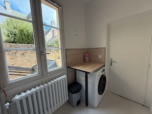 A laundry room with a washing machine, a small trash bin, a radiator, and a window with a view of a stone building and greenery. - Villa Ursuline: Charm and Comfort in the Heart of the City (Bayeux)