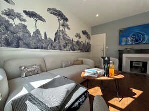 Ein Wohnzimmer mit einer Couch, einem Couchtisch mit Büchern und einer Flasche, einem Kamin und einem Wandgemälde.