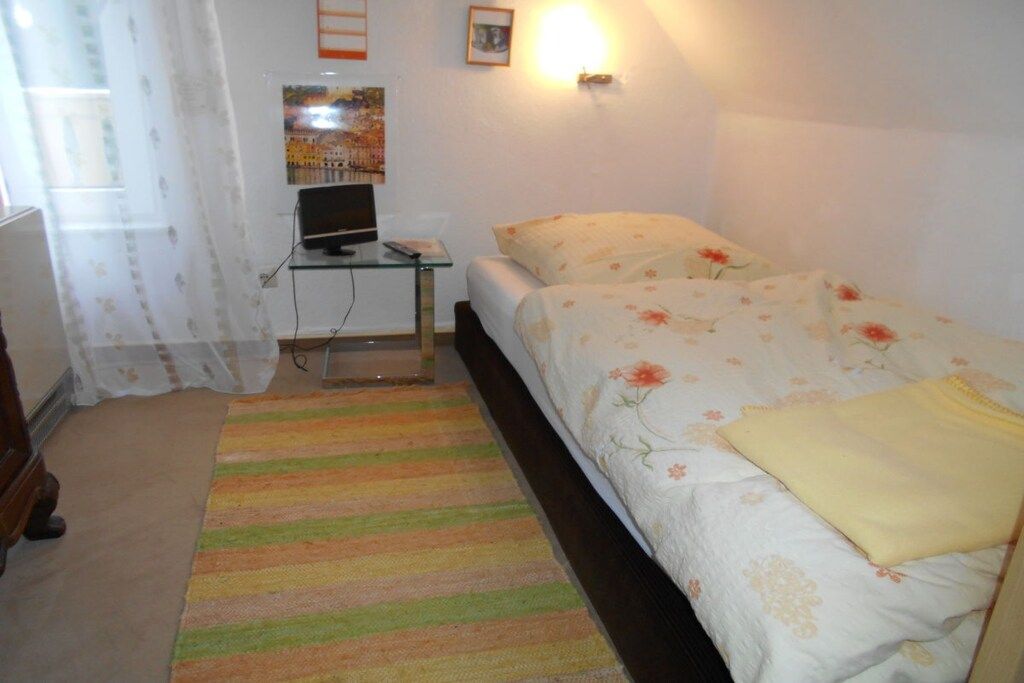 Ein Schlafzimmer mit einem Bett, einem Schreibtisch mit Computer, einer Lampe und einem Fenster mit Vorhängen.