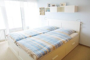 A double bed with a striped bedspread, a white headboard, and a bedside table with a drawer. There are wall-mounted shelves above the bed with framed pictures. - Kaiserhof with sea view - Spa Zone 1 (Norderney)