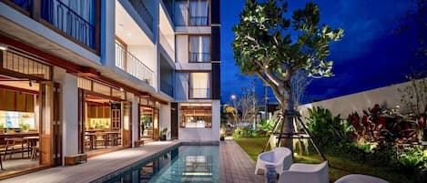 A hotel with a pool, outdoor seating, and a well-lit interior.