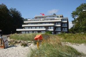 Ein mehrstöckiges Gebäude mit Flachdach und Balkonen, umgeben von Sand und Vegetation.