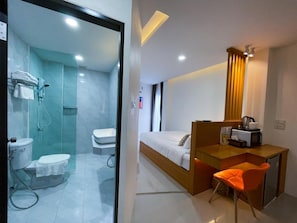 A modern hotel room with a glass shower enclosure, a separate toilet, a bed with a wooden headboard, a small desk with a chair, and a microwave.