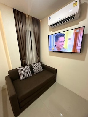 A room with a brown sofa, two checkered pillows, a flat-screen TV mounted on the wall, and an air conditioning unit above the sofa.