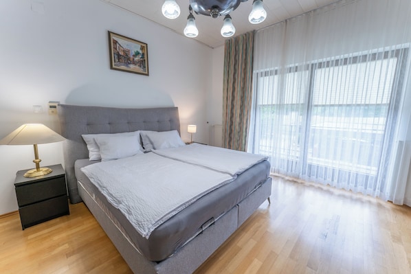 A modern bedroom with a large bed, bedside lamps, a nightstand, a painting on the wall, and sheer curtains on the window.