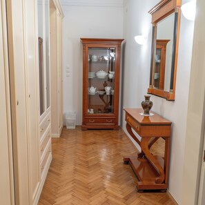 Een gang met houten vloer, een glazen kast met servies en een spiegel aan de wand.