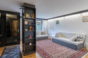 Un salon avec un canapé, un tapis à motifs, une bibliothèque et une fenêtre avec des stores.