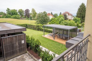 A rooftop terrace with a covered seating area, a grill, and a view of a green lawn and trees.